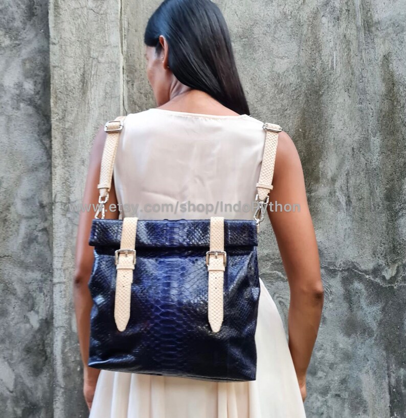 May include: A dark blue python leather backpack with cream-colored straps and buckles. The bag has a fold-over top and is worn by a person.