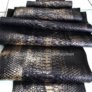 以下が含まれることがあります： 黒と金のパイソンレザーの革の束。革は、質感のある鱗模様をしています。革は折り畳まれ、互いに積み重ねられています。