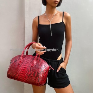 May include: A red snakeskin handbag with a top handle and a silver chain strap. The bag is being held by a person wearing a black bodysuit and black shorts.