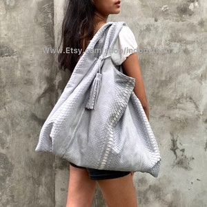May include: A large, light gray, slouchy tote bag made of snakeskin with a tassel. The bag is worn over the shoulder of a person standing in front of a gray wall.