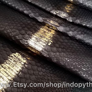 Op de afbeelding: Close-up van zwart pythonleer met gouden accenten. Het leer is in overlappende lagen gerangschikt, waardoor de textuur van de schubben zichtbaar is. De afbeelding bevat de tekst "www.Etsy.com/shop/indopython".