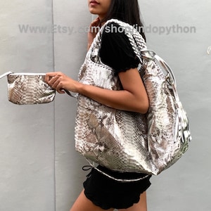 Op de afbeelding: Een zilveren slangenleren tote bag met een bijpassende slangenleren clutch. De tote bag heeft een lange band en een kwast. De clutch heeft een ritssluiting.