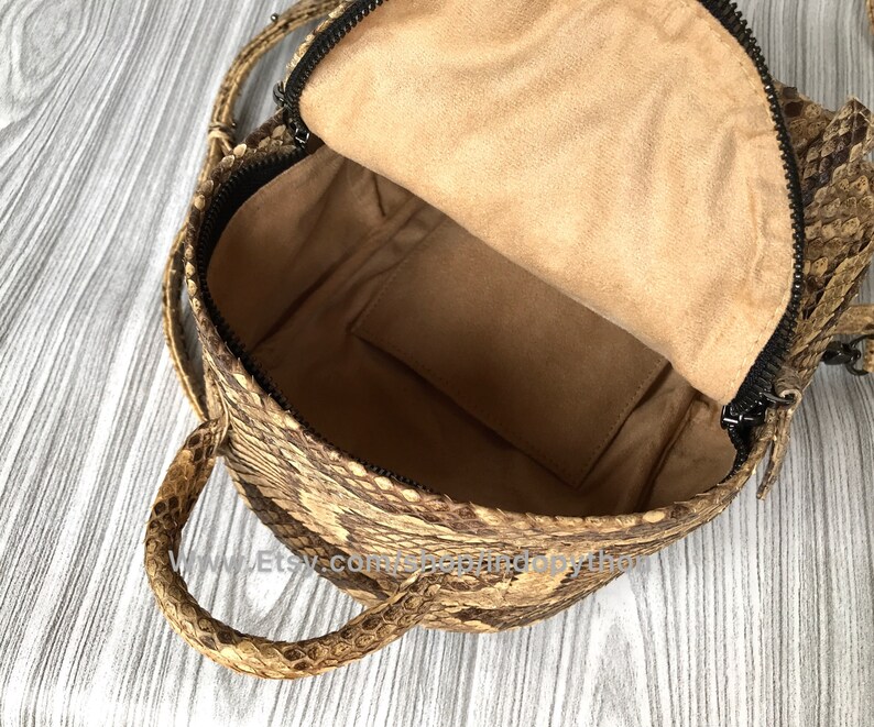 May include: Open view of a brown and tan snakeskin backpack with a black zipper. The interior is lined with tan fabric and includes a small pocket. The bag has a top handle and adjustable straps.