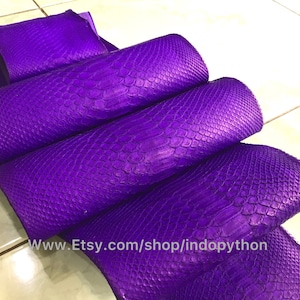 May include: Purple python skin, a natural material used for crafting leather goods. The skin is folded in a stack, showcasing its textured surface and vibrant colour.