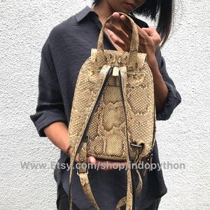 Puede incluir: Una peque&ntilde;a mochila de piel de serpiente beige con correas negras y cierre de cremallera. La mochila la lleva una persona que lleva una camisa negra.