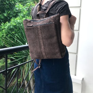 K&ouml;nnte beinhalten: Ein brauner Lederrucksack mit Schlangenhautmuster. Der Rucksack hat zwei Rei&szlig;verschlussf&auml;cher und verstellbare Tr&auml;ger.