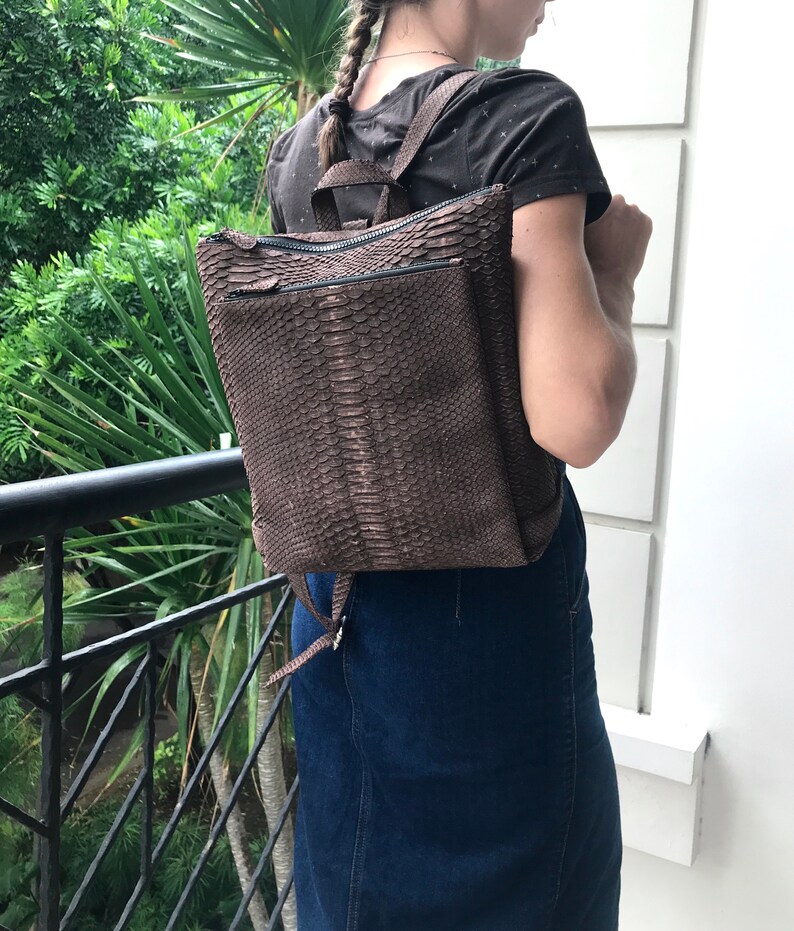 K&ouml;nnte beinhalten: Ein brauner Lederrucksack mit Schlangenhautmuster. Der Rucksack hat zwei Rei&szlig;verschlussf&auml;cher und verstellbare Tr&auml;ger.