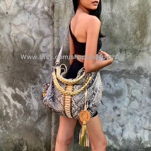 Może przedstawiać: Torebka skórzana ze skóry węża z złotym łańcuszkiem i frędzlem. Torebka ma biało-złoty wzór w paski i okrągły metalowy brelok.