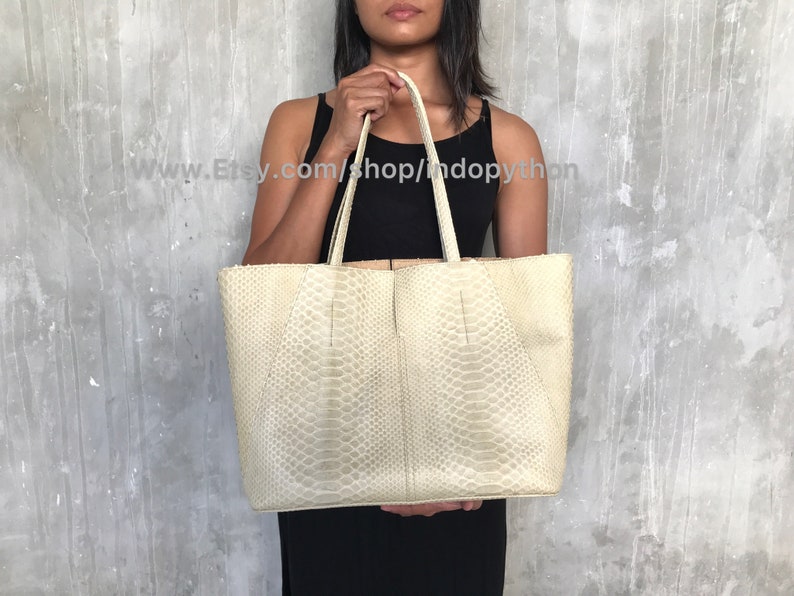 May include: A large, off-white tote bag made of snakeskin. The bag has a structured shape and two top handles.