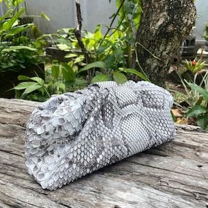 May include: A gray and white snakeskin clutch bag. The bag is made of genuine snakeskin with a scale pattern. The clutch is rectangular with a gathered top and is resting on a weathered wooden surface. The background features green foliage.