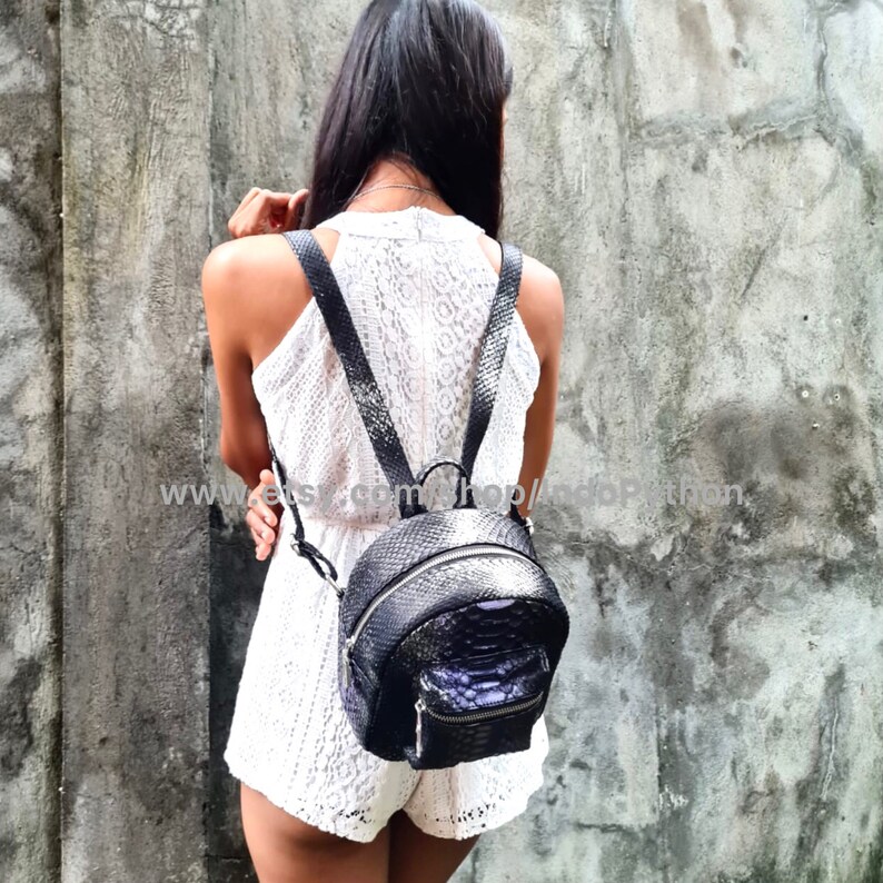 May include: A black leather backpack with a snakeskin pattern. The backpack has a round shape and a zipper closure. The straps are adjustable and the backpack has a small pocket on the front.