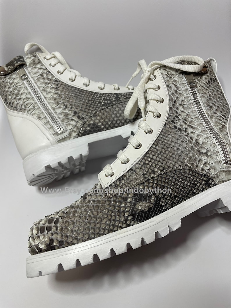 May include: White leather high-top sneakers with a snakeskin pattern and white laces. The sneakers have a zipper on the side and a white rubber sole.