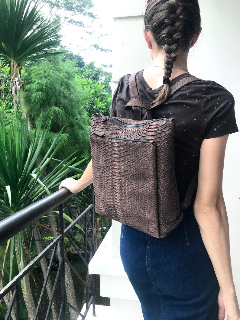 K&ouml;nnte beinhalten: Ein brauner Schlangenleder-Rucksack mit Rei&szlig;verschluss. Der Rucksack wird von einer Person mit langen braunen Haaren getragen.