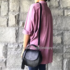 May include: A black leather handbag with a snakeskin pattern and a tassel. The bag has a shoulder strap and a top handle. The bag is being worn by a person wearing a pink shirt.
