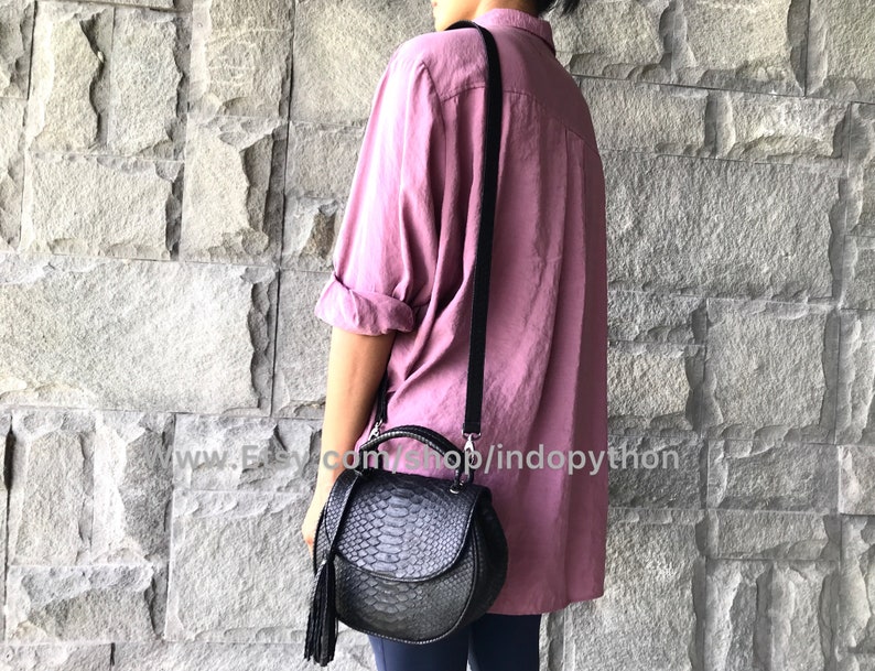 May include: A black leather handbag with a snakeskin pattern and a tassel. The bag has a shoulder strap and a top handle. The bag is being worn by a person wearing a pink shirt.