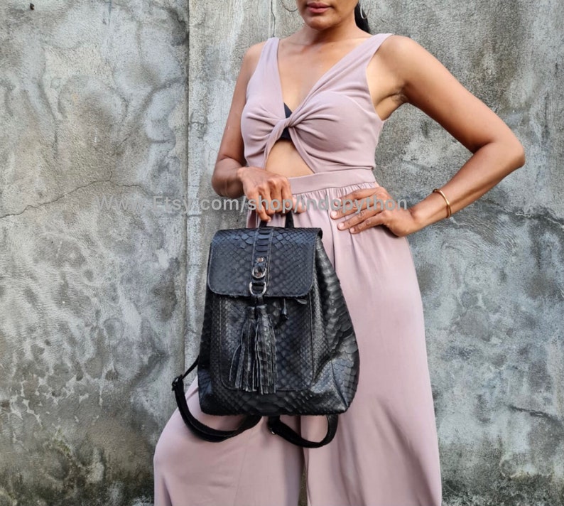 May include: A black leather backpack with a snakeskin pattern and a tassel. The backpack has a flap closure with a buckle and a zipped pocket on the front.