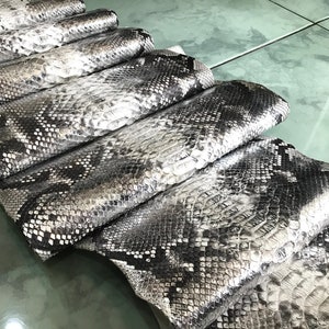 Op de afbeelding: Een stapel zilveren en zwarte pythonhuiden. De huiden zijn in een verspringend patroon gerangschikt, waarbij de bovenste huid het meest zichtbaar is. De huiden zijn glad en glanzend, en de schubben zijn duidelijk zichtbaar.