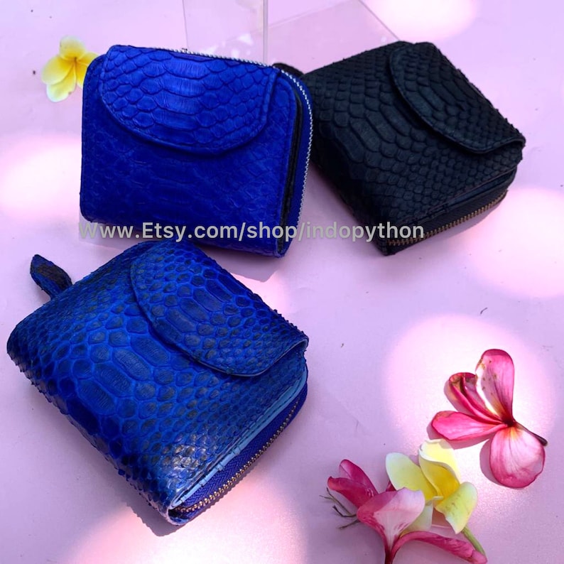 K&ouml;nnte beinhalten: Drei kleine blaue und schwarze Ledergeldb&ouml;rsen mit Rei&szlig;verschluss. Die Geldb&ouml;rsen sind aus Pythonleder gefertigt und haben ein einzigartiges, strukturiertes Muster. Die Geldb&ouml;rsen werden auf einer rosa Oberfl&auml;che mit Blumen pr&auml;sentiert.