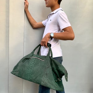 May include: A green snakeskin leather travel bag with a zipper closure and a silver buckle. The bag is being held by a person wearing a white polo shirt and blue jeans.