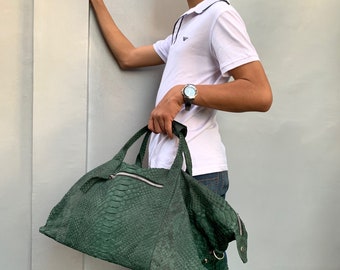 May include: A green snakeskin leather travel bag with a zipper closure and a silver buckle. The bag is being held by a person wearing a white polo shirt and blue jeans.