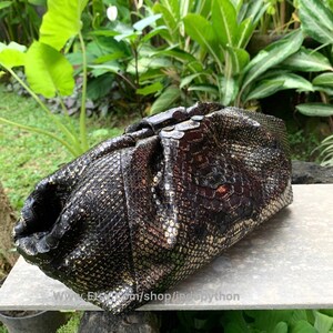 K&ouml;nnte beinhalten: Eine schwarze Clutch aus Pythonleder mit goldenen Akzenten und einem gerafften Design. Die Tasche ist aus echtem Pythonleder gefertigt und hat eine einzigartige, strukturierte Optik.