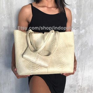 May include: A large, off-white tote bag made of python skin. The bag has two top handles and a smaller, matching python skin pouch attached to the front.