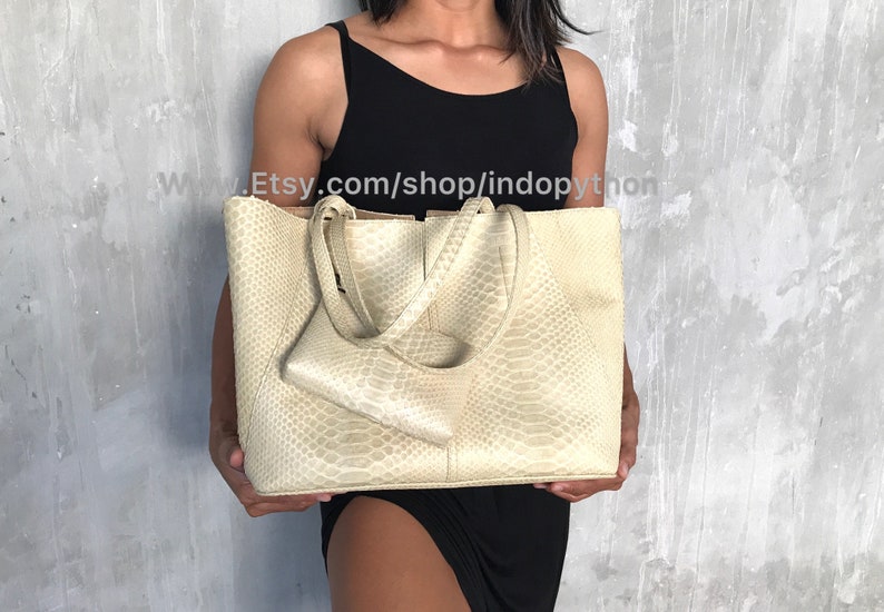 May include: A large, off-white tote bag made of python skin. The bag has two top handles and a smaller, matching python skin pouch attached to the front.