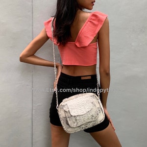 May include: A white snakeskin crossbody bag with a silver chain strap. The bag has a flap closure and a small pocket on the front. The bag is being worn by a person wearing a pink crop top and black shorts.