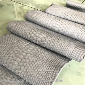 Op de afbeelding: Grijze python lederhuid, gevouwen en gepresenteerd op een lichtgrijze betegelde ondergrond. Het leer heeft een getextureerd, schubbig patroon.