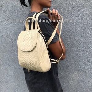 May include: A cream-colored snakeskin backpack with black straps. The backpack has a flap closure and a zippered pocket on the front.