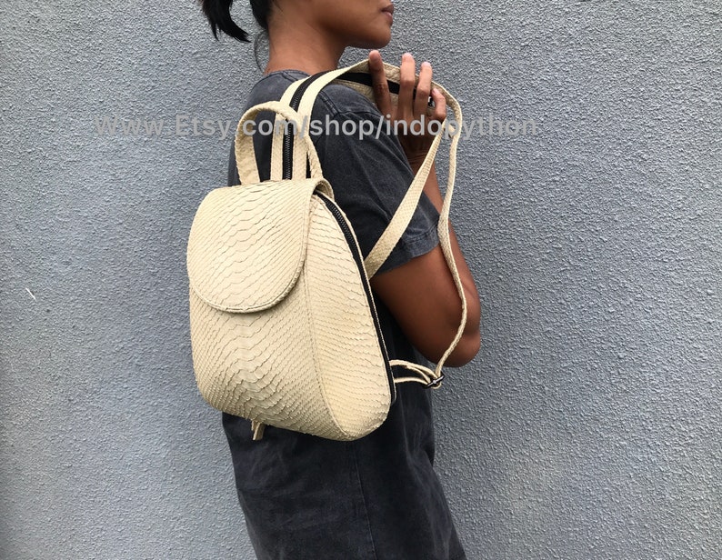 May include: A cream-colored snakeskin backpack with black straps. The backpack has a flap closure and a zippered pocket on the front.