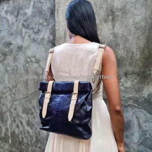 May include: A blue leather backpack with a fold-over top and buckle closures. The straps are a light beige color. The backpack is being worn by a person.