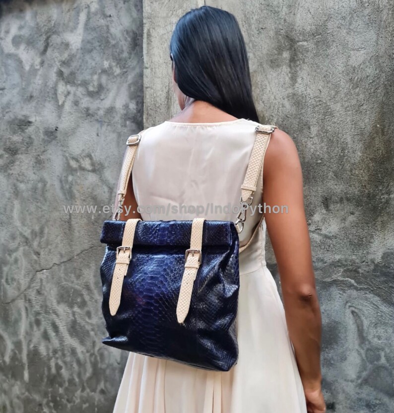 May include: A blue leather backpack with a fold-over top and buckle closures. The straps are a light beige color. The backpack is being worn by a person.