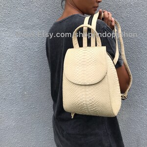 May include: A small, off-white leather backpack with a snakeskin pattern. The backpack has a flap closure and a drawstring closure at the bottom. The straps are adjustable and have a black zipper detail.