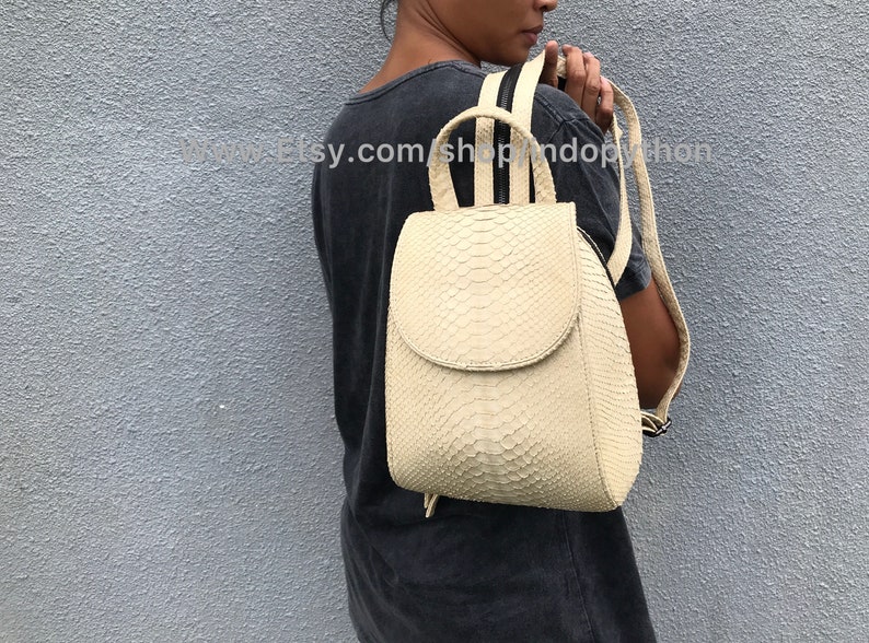 May include: A small, off-white leather backpack with a snakeskin pattern. The backpack has a flap closure and a drawstring closure at the bottom. The straps are adjustable and have a black zipper detail.