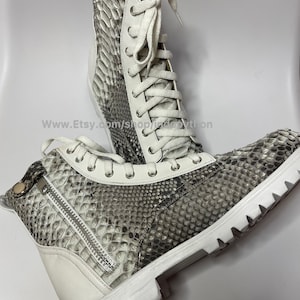 May include: White leather boots with a snakeskin pattern and white laces. The boots have a zipper on the side and a thick white sole.