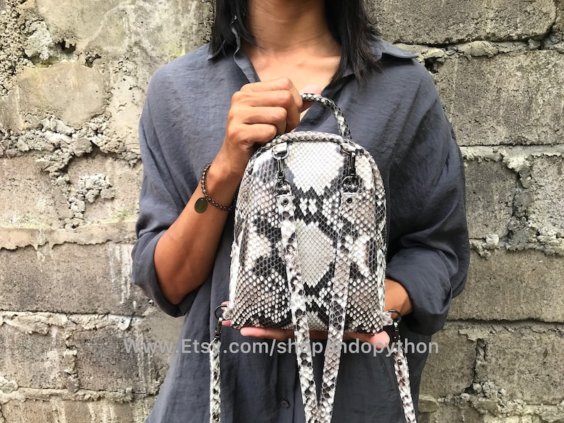 K&ouml;nnte beinhalten: Ein kleiner, wei&szlig;er und schwarzer Schlangenleder-Rucksack mit verstellbaren Tr&auml;gern. Der Rucksack wird von einer Person in einem grauen Shirt getragen.