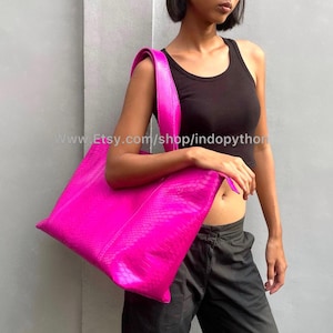 Puede incluir: Un bolso tote fucsia vibrante con un patrón texturizado similar a la piel de serpiente. El bolso tiene forma rectangular y una correa ancha para el hombro. El bolso lo lleva una persona que viste una camiseta sin mangas negra y pantalones negros.