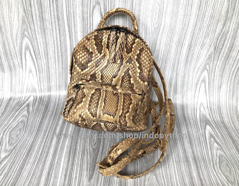 May include: A brown and tan snakeskin backpack with a top handle and adjustable straps. The bag has a front zippered pocket and a small loop for hanging. The snakeskin pattern is visible across the entire bag.