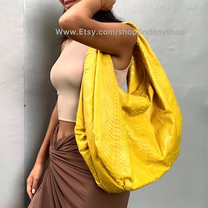 Può includere: Una borsa a mano in pelle di pitone giallo brillante con uno stile ampio e hobo. La borsa è indossata sulla spalla di una persona che indossa una gonna marrone e una canotta beige.
