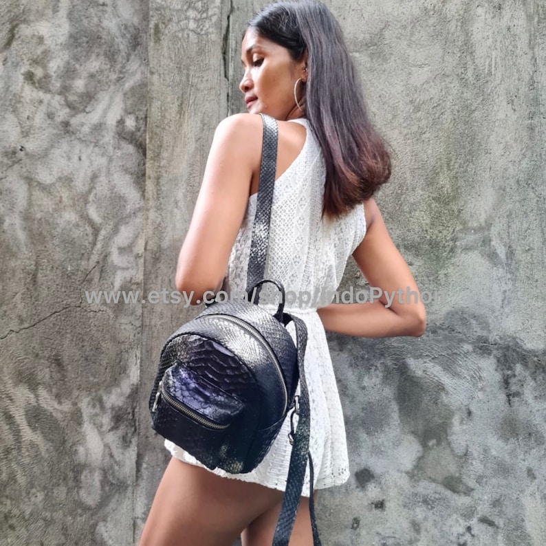 May include: A black leather backpack with a snakeskin pattern. The backpack has a zipper closure and a small pocket on the front. The backpack is being worn by a person.