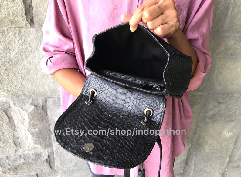 May include: Black leather handbag with a snakeskin pattern. The bag is open and the inside is lined with black fabric. The bag has a zipper closure and a drawstring closure.