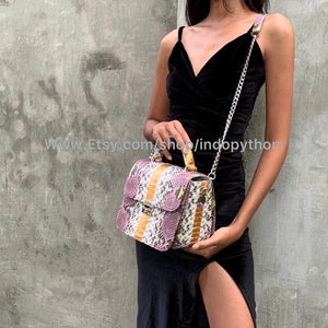 May include: A small, rectangular handbag with a snakeskin pattern in shades of purple, yellow, and white. The bag has a top handle and a silver chain strap. The bag is being held by a person wearing a black dress.