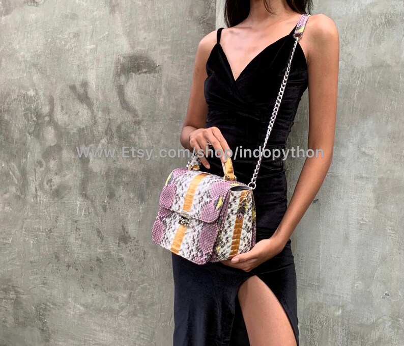May include: A small, rectangular handbag with a snakeskin pattern in shades of purple, yellow, and white. The bag has a top handle and a silver chain strap. The bag is being held by a person wearing a black dress.