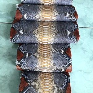 May include: Several rolls of python leather, displaying a natural pattern of grey, brown, and tan scales. The leather has a textured appearance, with a repeating pattern. The edges are a darker brown.