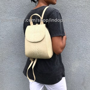 May include: A cream-colored snakeskin backpack with a flap closure and adjustable straps. The backpack is worn by a person against a gray wall.