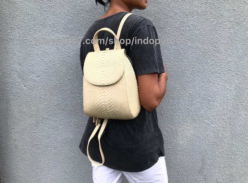 May include: A cream-colored snakeskin backpack with a flap closure and adjustable straps. The backpack is worn by a person against a gray wall.