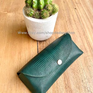 Op de afbeelding: Een donkergroene, getextureerde leren brillenkoker met een zilveren knoopsluiting. De koker is rechthoekig met een gebogen bovenkant. Een kleine cactus in een witte pot staat op de achtergrond. De Etsy shop URL is zichtbaar.