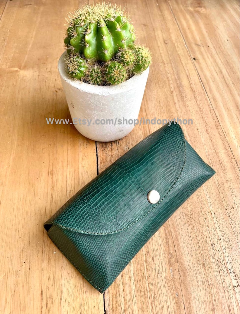 Op de afbeelding: Een donkergroene, getextureerde leren brillenkoker met een zilveren knoopsluiting. De koker is rechthoekig met een gebogen bovenkant. Een kleine cactus in een witte pot staat op de achtergrond. De Etsy shop URL is zichtbaar.