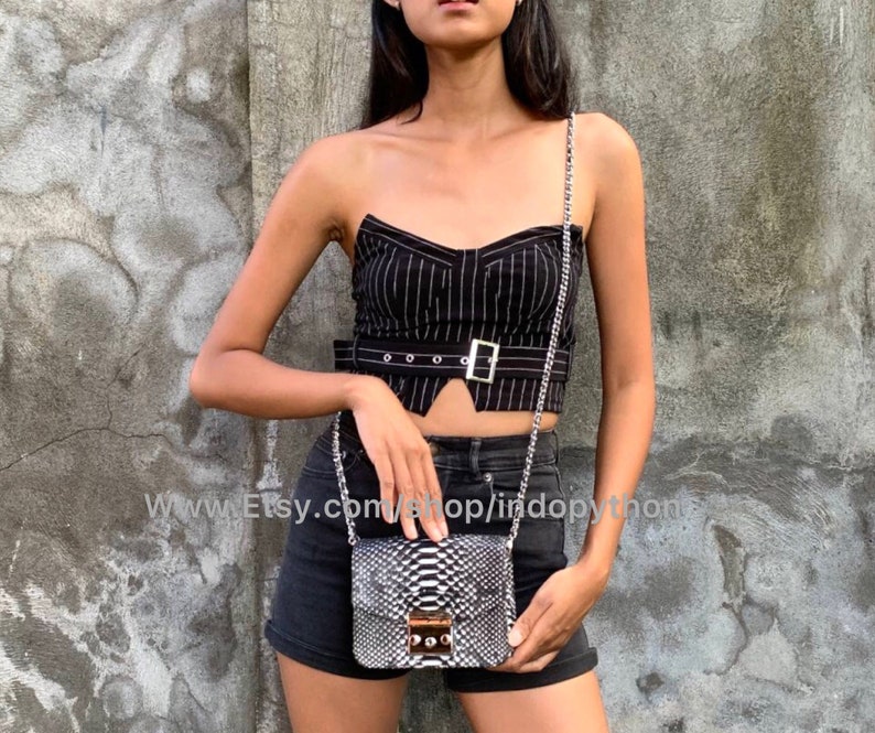 May include: A black and white snakeskin handbag with a silver chain strap. The bag has a gold clasp and is being worn by a person wearing a black and white striped top and black shorts.